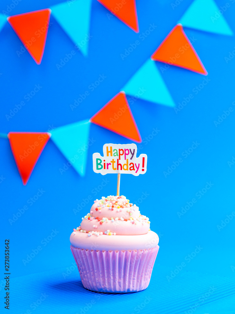 Pink cupcake with Happy Birthday text and holiday flags on a blue background. Vertical shot