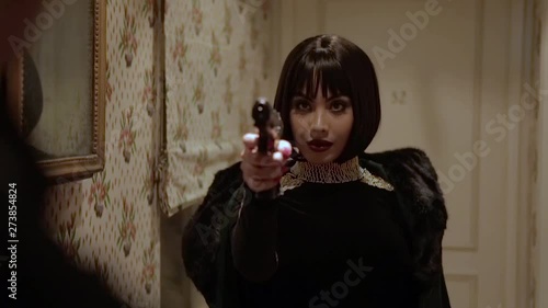 Sexy brunette Arab woman in tight black dress with jewels and big pearl necklace pointing a gun on her husband who cheated on her in a retro hotel with walls adorned with floral tapestry. Slow motion.