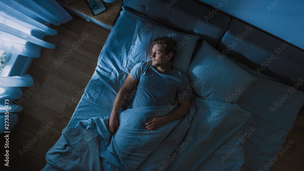 © Gorodenkoff - Top View of Handsome Young Man Sleeping Cozily on a Bed in His Bedroom at Night. Blue Nightly Colors with Cold Weak Lamppost Light Shining Through the Window. © Gorodenkoff - Top View of Handsome Young Man Sleeping Cozily on a Bed in His Bedroom at Night. Blue Nightly Colors with Cold Weak Lamppost Light Shining Through the Window.