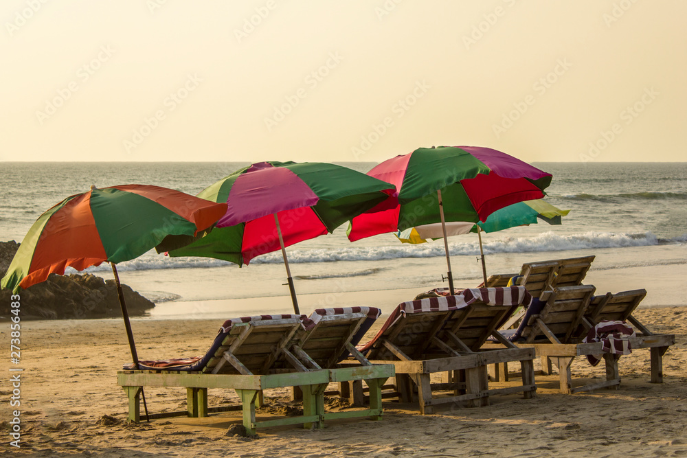 empty wooden beach loungers with striped mattresses under bright large sun umbrellas on the sand against the background of the ocean with sea waves under the gray evening sky