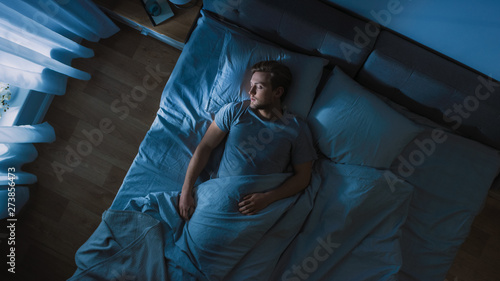 Top View of Handsome Young Man Sleeping Cozily on a Bed in His Bedroom at Night. Blue Nightly Colors with Cold Weak Lamppost Light Shining Through the Window.