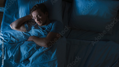 Top View of Handsome Young Man Sleeping Cozily on a Bed in His Bedroom at Night. Blue Nightly Colors with Cold Weak Lamppost Light Shining Through the Window. photo