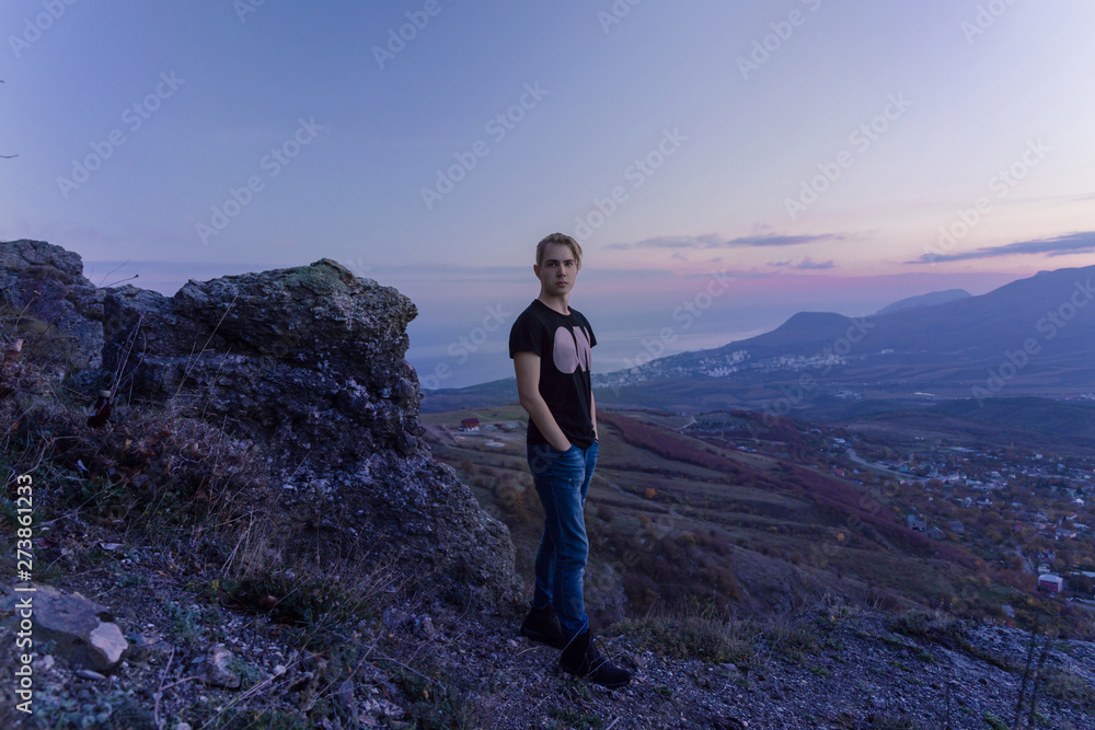 Portrait in growth. Young attractive man standing on the top of mountains looks to the camera, violet sunset.