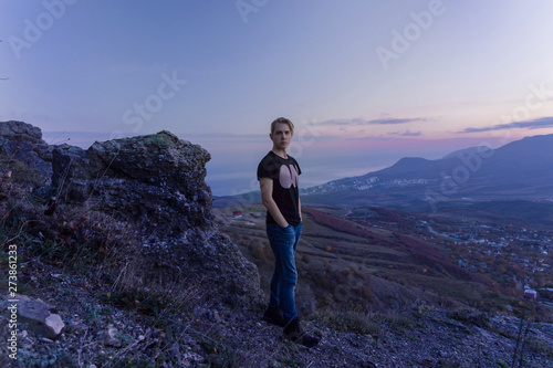 Portrait in growth. Young attractive man standing on the top of mountains looks to the camera, violet sunset.
