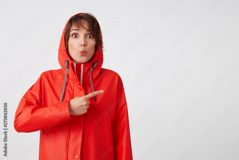 Wow! Cool news! Amazed short haired lady in red rain coat wants to draw your attention, points fingers to the copy space. Standing over white background. Wow! Cool news! Amazed short haired lady in red rain coat wants to draw your attention, points fingers to the copy space. Standing over white background.
