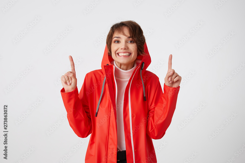 Cute positive short haired girl in red rain coat, broadly smiles, looks at the camera, wants to draw your attention to the copy space above her head, points fingers up. Standing over white background. Cute positive short haired girl in red rain coat, broadly smiles, looks at the camera, wants to draw your attention to the copy space above her head, points fingers up. Standing over white background.