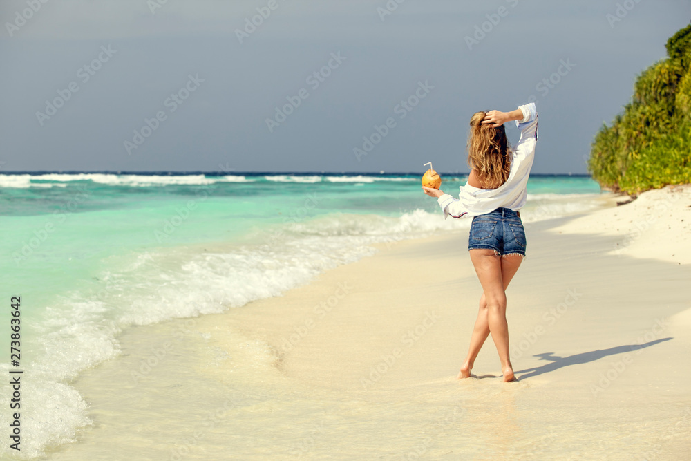 Blonde girl in denim shorts with fresh coconut in hand, on beach ocean in Maldives. Summer vacation travel. Banner panorama landscape. Blonde girl in denim shorts with fresh coconut in hand, on beach ocean in Maldives. Summer vacation travel. Banner panorama landscape.