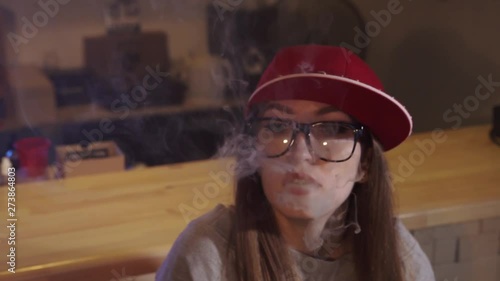 Young pretty woman in red cap smoke an electronic cigarette at the vape shop. Closeup. Slow motion. photo