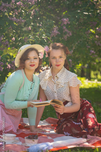 two pretty girls friends sitting on the red blanket on the green grass and have summer picnic. happy woman having rest and fun on their vacantions on the nature. photo