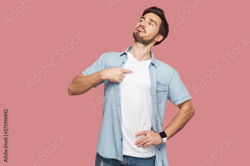 This is me. Portrait of proud haughty handsome bearded young man in blue casual style shirt standing, looking away and pointing himself. indoor studio shot, isolated on pink background.