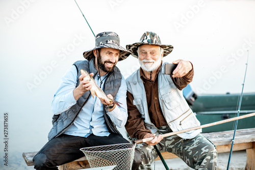 Grandfather with son having fun, making selfie photo together with freshly caught fish on the lake early in the morning