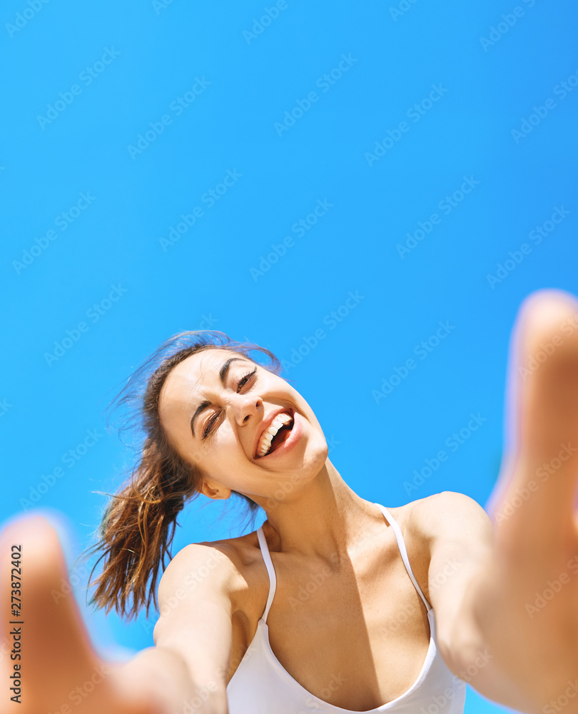 Portrait of beautiful happy woman taking a selfie on the camera phone with blue sky on background. Young woman having fun smiling winking and laughing against blue clear sky. Summertime and leisure