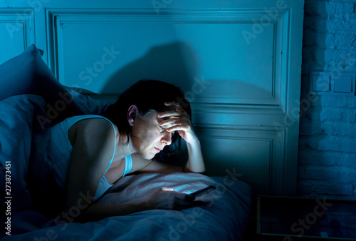 Canvastavla young beautiful woman lying bored in bed using smart phone late at night in a da