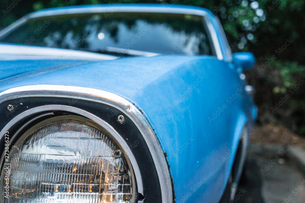 Close up of vintage blue car's old headlamp. Parked and surrounded by trees.