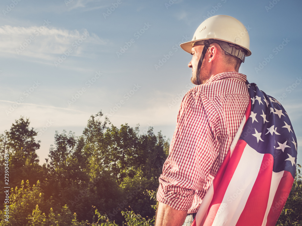 Young engineer, holding white hardhat and an American Flag in the park against the backdrop of green trees and the setting sun, looking into the distance. Close-up. Concept of labor and employment