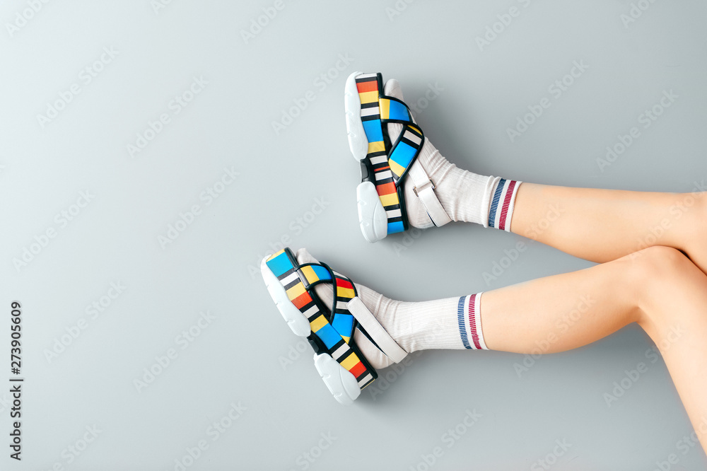 Beautiful female legs in white trendy socks posing in colorful fashionable  high wedge leather sandals on gray background. Asian anime style concept.  Womens legs wearing high sole summer stylish shoes. Stock Photo |
