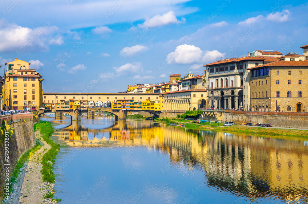 Ponte Vecchio bridge with colourful buildings houses over Arno River blue reflecting water and embankment promenade in historical centre of Florence city, blue sky white clouds, Tuscany, Italy