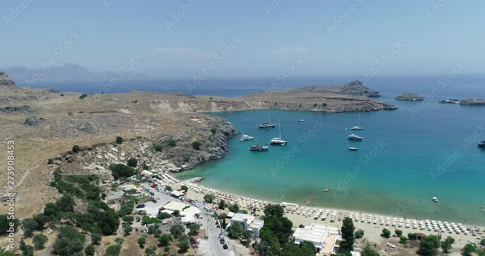 Lindos bay. Rhodes island. Greece. Aerial view from Lindos village. Idyllic sea coast landscape with blue sky. Tourist ships and boats. White beach umbrellas. Lindos bay. Rhodes island. Greece. Aerial view from Lindos village. Idyllic sea coast landscape with blue sky. Tourist ships and boats. White beach umbrellas.