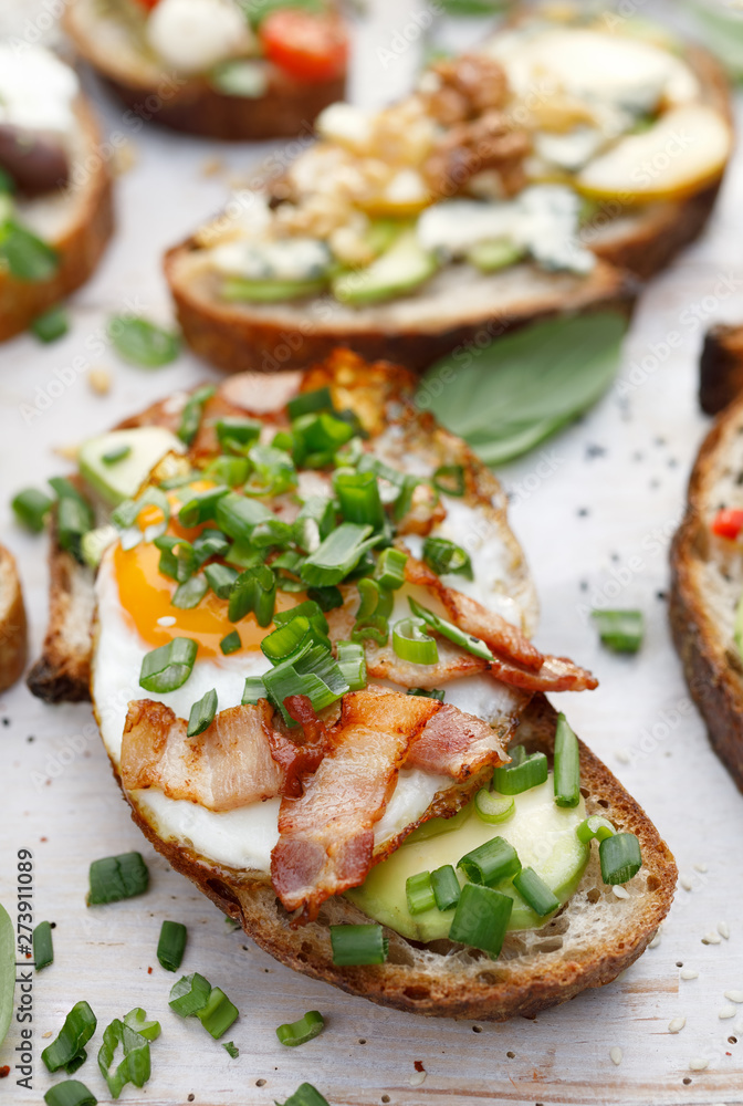 Open sandwich made of slices of sourdough bread with addition of avocado, egg, bacon, fresh scallion on a wooden white table, close-up. 