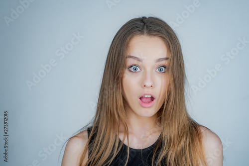 Young attractive girl with surprise curiosity emotion looking at camera and open lips. Open mouth. Portrait of cheerful beautiful woman over gray background. People emotions concept.