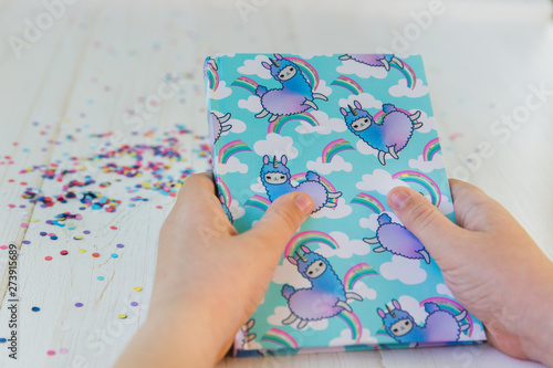 Hands of a girl holding notebook with llama unicorn and rainbow on white wooden background with confetti. Idea of Girly Desk table