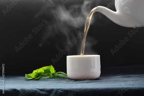 cup of tea with mint and pouring tea white teapot and smoke black background