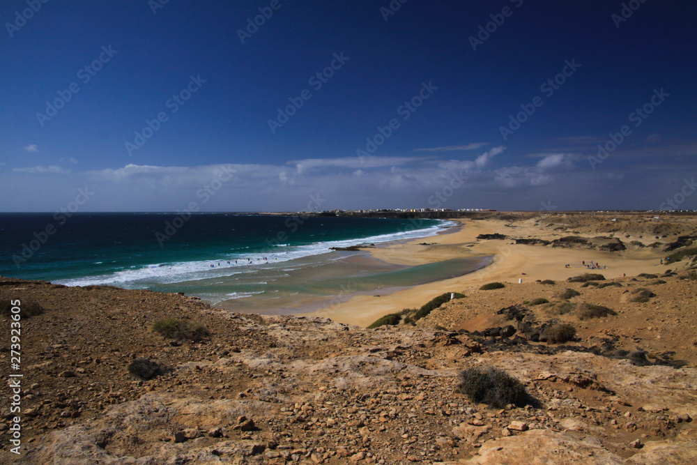 View over sand dunes with green ocean on (Playa del Aljibe) on white village on steep cliff (El Cotillo) - North Fuerteventura