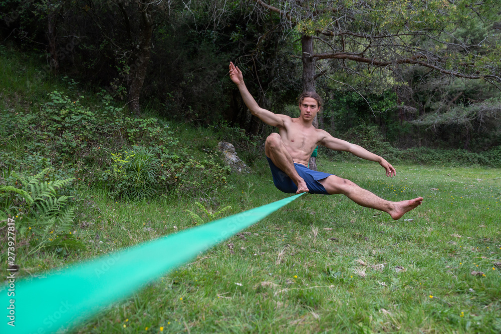 Active young man with naked torso balancing on slackline in green field on summertime