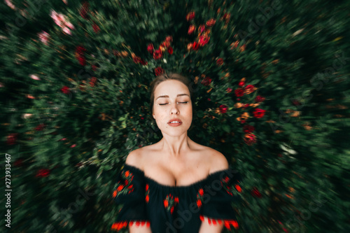 Adorable fabulous young girl in embroidered dress with eyes closed dreaming at rose bushes on background. photo