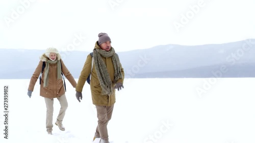 Full shot of fit 60-something Caucasian couple hiking in ankle-deep snow over frozen lake on cloudy winter day, with husband turning to his wife, talking and laughing, and wife responding with smile