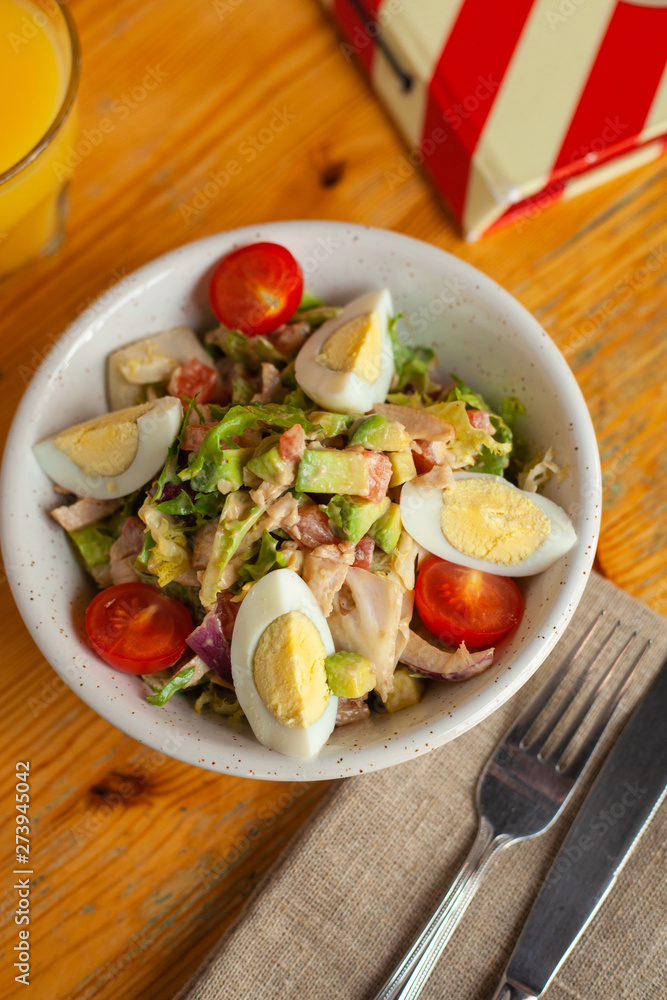Traditional american cobb salad with chopped avocado, tomatoes, hard boiled egg, roasted chicken and greens for a healthy lunch, served on wooden table with soft light