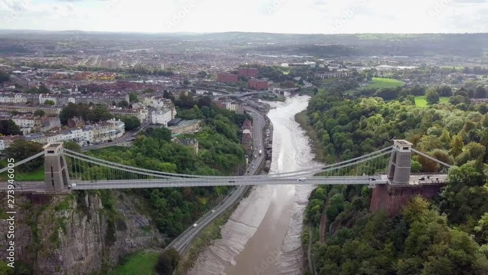 A drone fly over shot of Clifton Suspension Bridge above the Avon River, and highway, in Bristol, England, on a clear day. A reveal of Bristol's skyline of its buildings and historic landscape. A drone fly over shot of Clifton Suspension Bridge above the Avon River, and highway, in Bristol, England, on a clear day. A reveal of Bristol's skyline of its buildings and historic landscape.