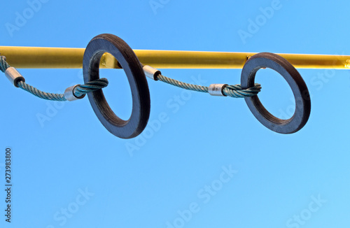Brown wooden gymnastic rings are suspended by blue twisted ropes with compressed metal hoops from yellow crossbar on Playground against blue sky. Concept: Adolescent and children activity and sports
