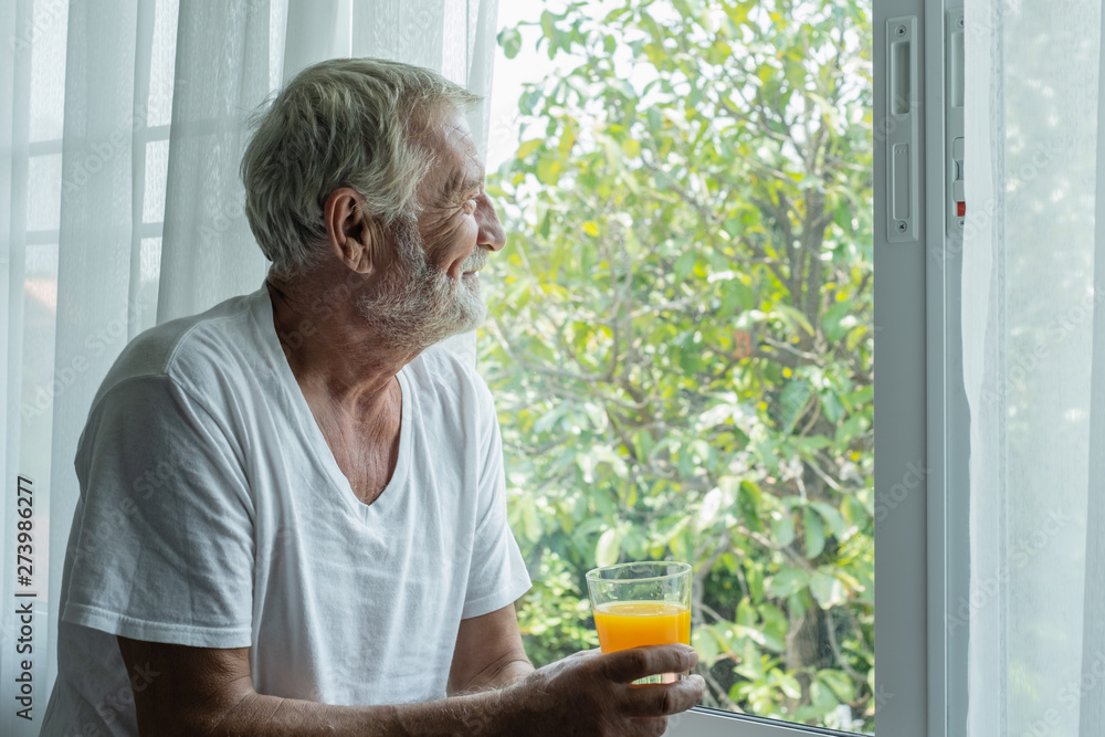 senior man hold glass of orange juice and thinking and looking outside of window in white room senior man hold glass of orange juice and thinking and looking outside of window in white room