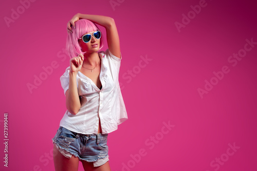 Concept of merry summer mood, relaxation and beauty. Close-up of a beautiful woman in a pink wig  in short attractive jeans, white shirt, sunglasses   posing on a pink background