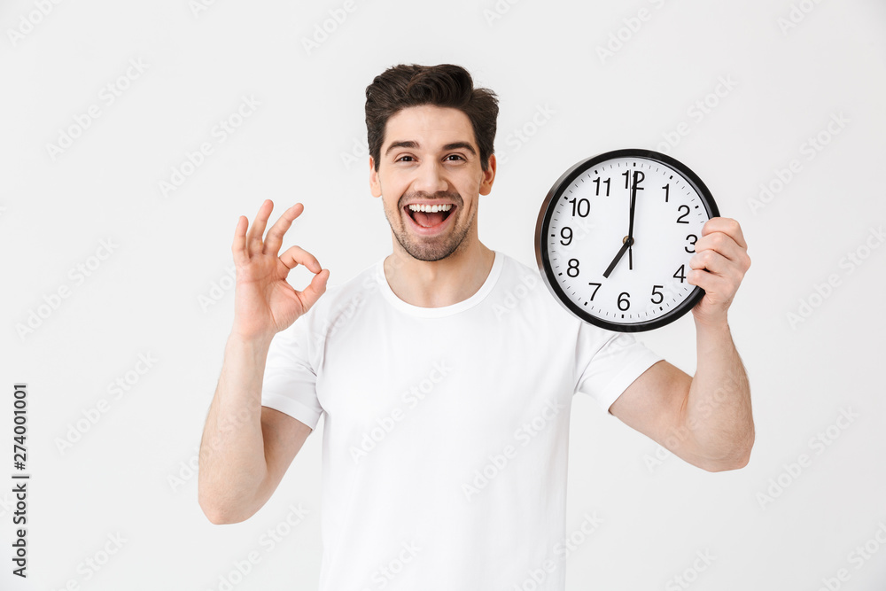 Shocked excited happy young man posing isolated over white wall background holding clock. Shocked excited happy young man posing isolated over white wall background holding clock.