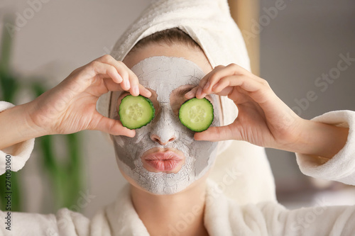 Young woman with clay mask and cucumber slices at home