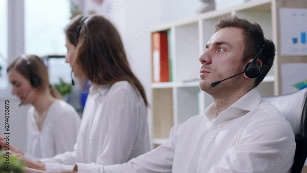 Focus on male call center worker accompanied by his female team. Customer support operator at work. Young female employees working with a headset and customers, male operator takes notes in notebook Focus on male call center worker accompanied by his female team. Customer support operator at work. Young female employees working with a headset and customers, male operator takes notes in notebook