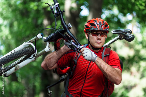 Positive sporty male holds mountain bicycle on a shoulder. Outdoor portrait of a rider in red sportswear, sunglasses and helmet