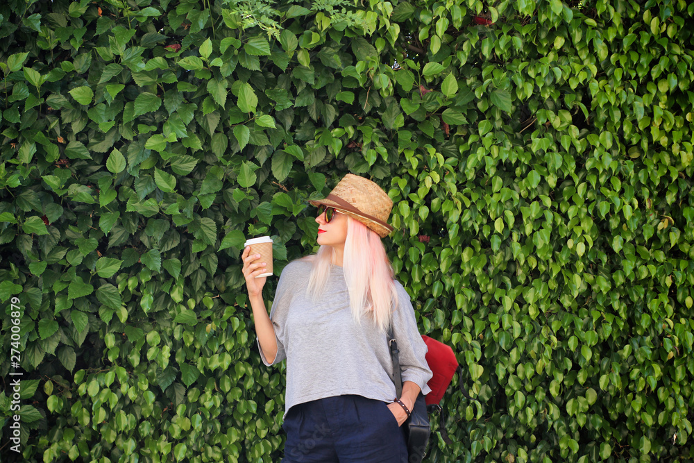 Young woman with pink blond hair, sunglasses, straw hat, paper cup of coffee, red lips and leather backpack on green wall background texture. 