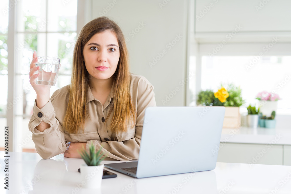 Beautiful young woman working with computer takes a break to drink glass of water with a confident expression on smart face thinking serious Beautiful young woman working with computer takes a break to drink glass of water with a confident expression on smart face thinking serious