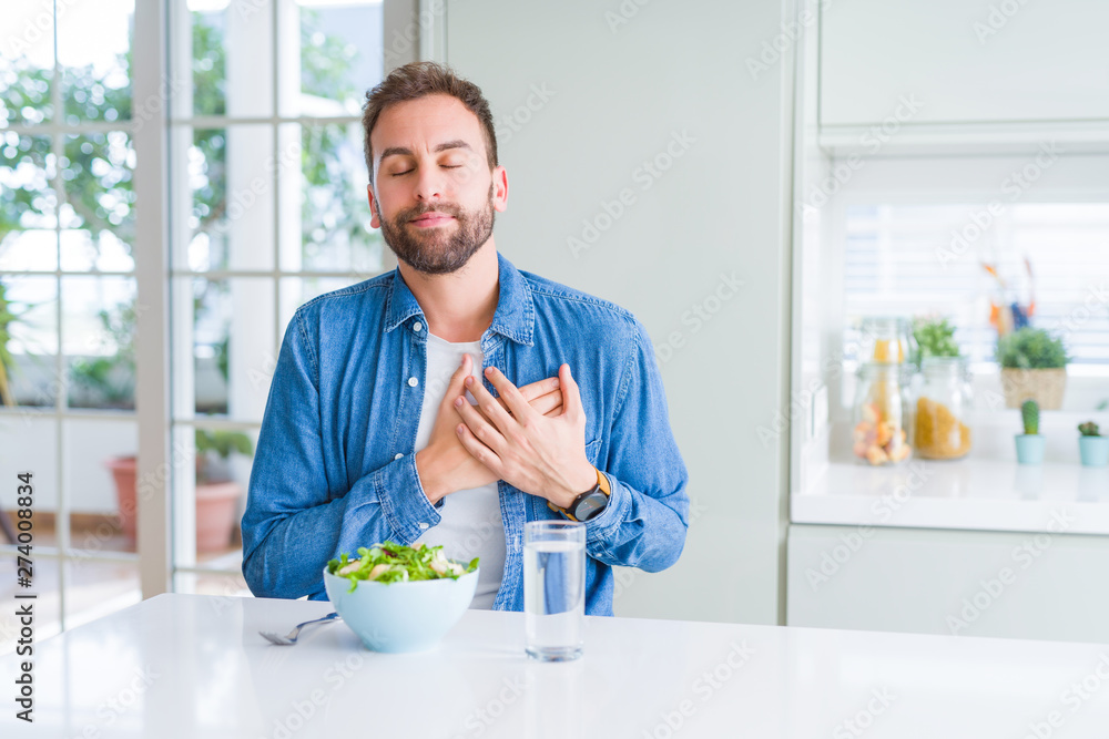 Handsome man eating fresh healthy salad smiling with hands on chest with closed eyes and grateful gesture on face. Health concept.