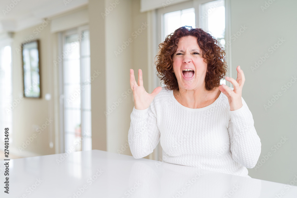 Beautiful senior woman wearing white sweater at home crazy and mad shouting and yelling with aggressive expression and arms raised. Frustration concept. Beautiful senior woman wearing white sweater at home crazy and mad shouting and yelling with aggressive expression and arms raised. Frustration concept.