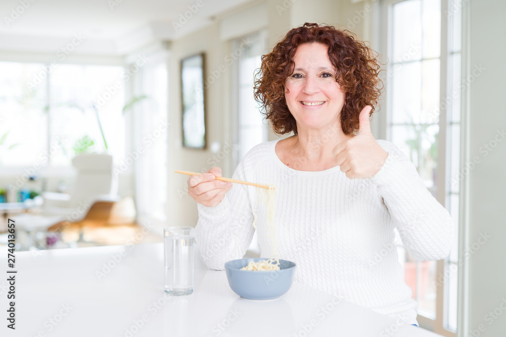 Senior woman eating asian noodles using chopsticks happy with big smile doing ok sign, thumb up with fingers, excellent sign Senior woman eating asian noodles using chopsticks happy with big smile doing ok sign, thumb up with fingers, excellent sign