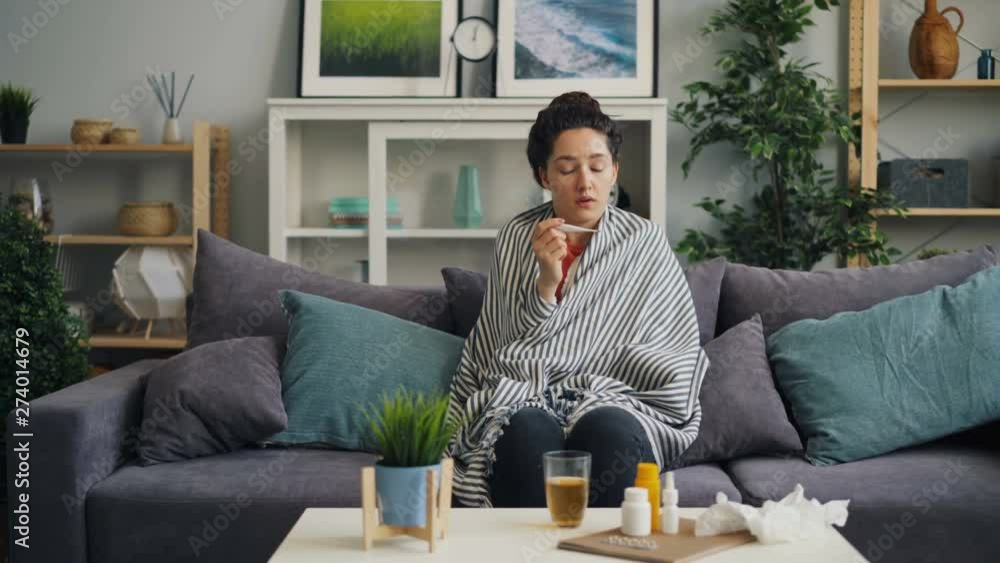 Sick female student taking temperature then drinking medicine at home suffering from cold alone sitting on couch wrapped in plaid. Medical problems and fever concept. Sick female student taking temperature then drinking medicine at home suffering from cold alone sitting on couch wrapped in plaid. Medical problems and fever concept.