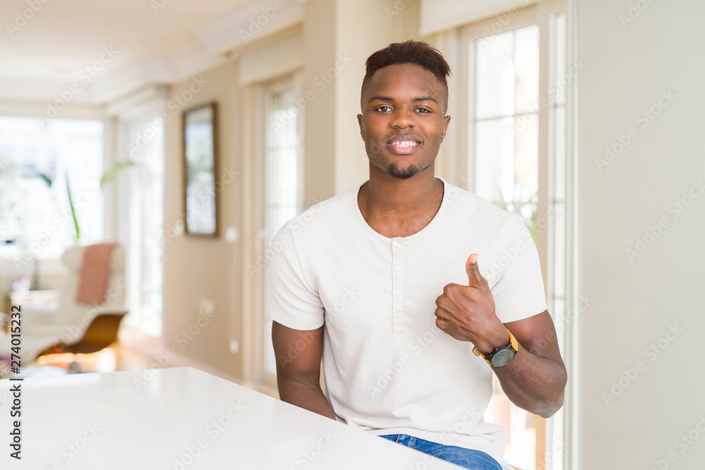 Handsome african american man on white table at home doing happy thumbs up gesture with hand. Approving expression looking at the camera with showing success. Handsome african american man on white table at home doing happy thumbs up gesture with hand. Approving expression looking at the camera with showing success.