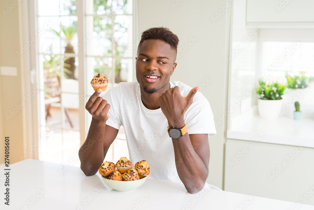 African american man eating chocolate chips muffin pointing and showing with thumb up to the side with happy face smiling African american man eating chocolate chips muffin pointing and showing with thumb up to the side with happy face smiling