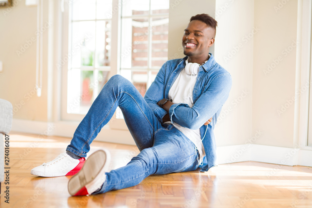 African american man wearing headphones listening to music sitting on the floor happy face smiling with crossed arms looking at the camera. Positive person.