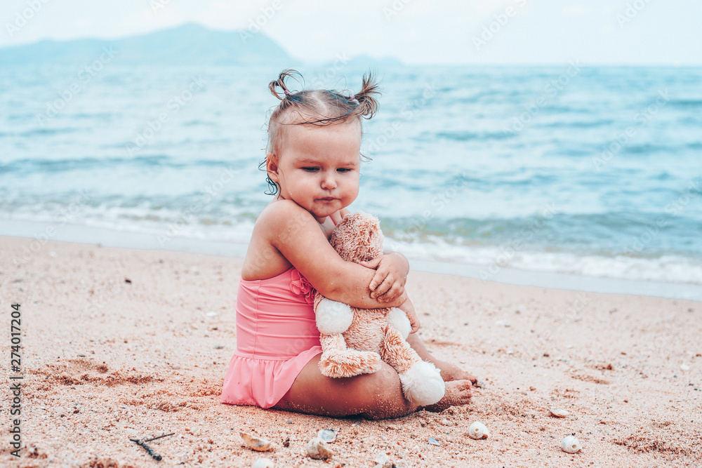 pensive little girl hugging teddy bear and looking away while sitting on seashore. pensive little girl hugging teddy bear and looking away while sitting on seashore.