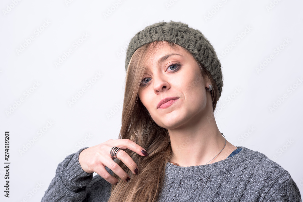 Confident funky hipster girl with knitted gray cap brushing hair looking at camera on light gray background. Confident funky hipster girl with knitted gray cap brushing hair looking at camera on light gray background.